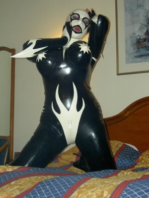 Fetish model Darkwing Zero poses on a hotel room bed in latex clothing