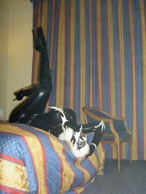 Fetish model Darkwing Zero poses on a hotel room bed in latex clothing