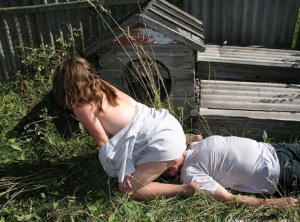 Caucasian girl straddles a man's face outside of a doghouse in a yard 21091960