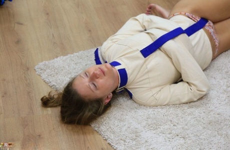 Caucasian girl is hogtied on a rug while wearing a straitjacket